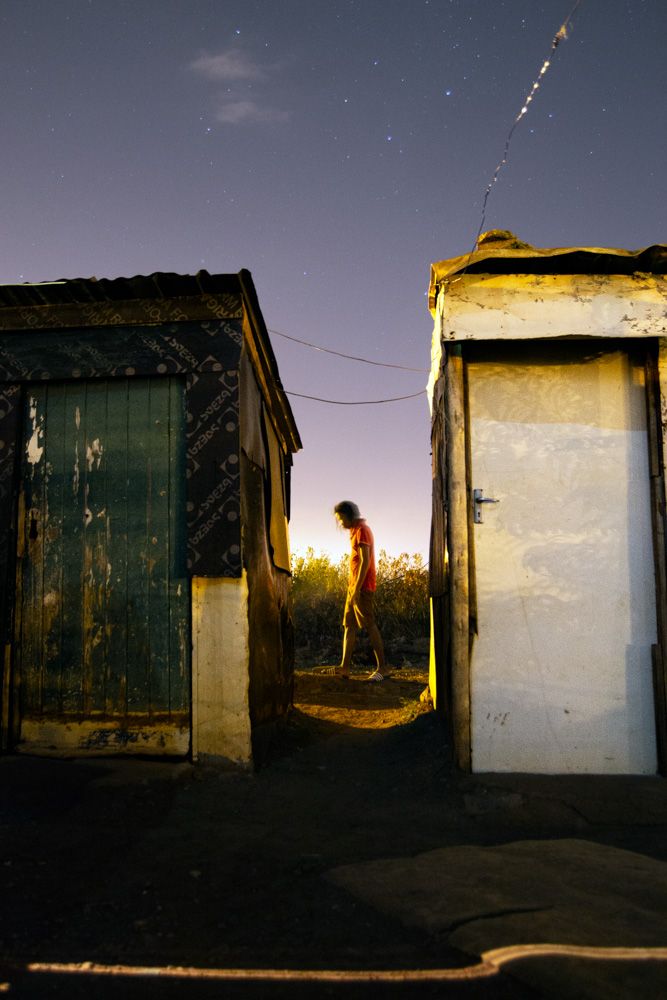 A conceptual nocturnal photograph titled "uMntu" by Sibusiso Bheka, exhibited at Afronova Gallery. The image uses cinematic lighting and silhouettes to document township life in Thokoza, focusing on atmospheric resilience and human presence.