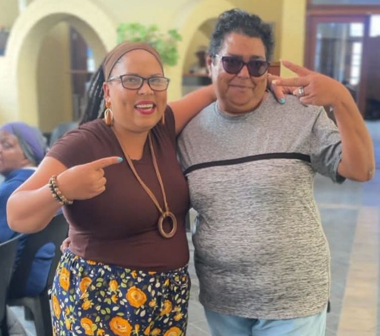 A candid photograph of community leader Janine "Blaq Pearl" Overmeyer engaged in a dialogue session with a participant at the Love Jam Session in Alliance Française, Mitchell's Plain. The setting is intimate and collaborative, reflecting the theme of connection.